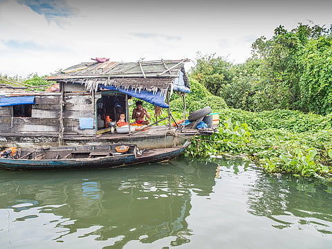 A rustic wooden boat on a calm river, covered with tarp and foliage. Two people handle daily tasks inside, surrounded by lush green vegetation.
