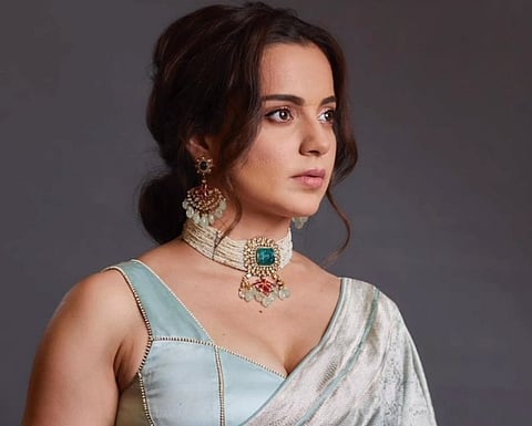 Image of Kangana ranaut in a blue saree and neck piece with an green emerald on it. In the image she is looking sideways aways from the camera.
