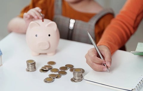 Parent writes in a notebook beside a child and pink piggy bank, with coin stacks on the table, illustrating family saving and financial education.