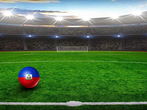 A Haiti-flag soccer ball rests on a bright green football field in a floodlit stadium, with a goal, packed stands, and blue sky beyond.