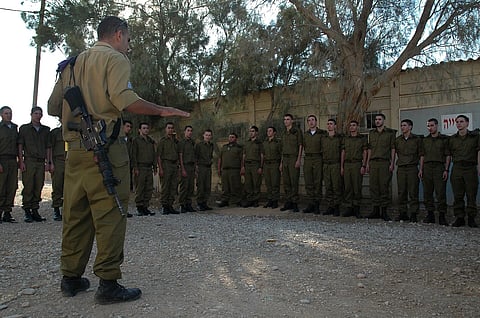 A uniformed Israeli soldier addresses a formation of troops outdoors, with rifles visible, under trees beside a concrete wall; the mood is serious.