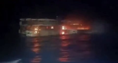 At night, a dimly lit vessel burns on dark water, with orange flames, smoke, and shimmering red reflections creating an ominous mood.