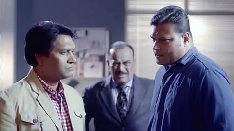 Three men stand in an office during a tense conversation: a casually dressed man faces a stern man in blue, while an older suited man watches.