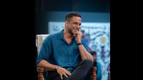 एक आदमी कुर्सी पर बैठा मुस्कुराते हुए नजर आ रहा है।