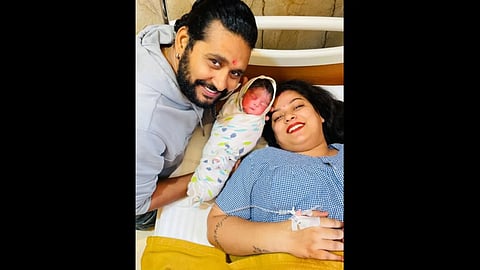 अस्पताल में महिला बिस्तर पर मुस्कुराती बैठी, पुरुष हाथ में नवजात शिशु लिए हुए, परिवार का खुशी भरा पल|