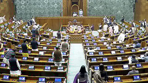 एक बड़े विधानसभा सदन का दृश्य, जहाँ कई सदस्य अपनी सीटों के पास खड़े हैं। सामने की ओर मंच पर कुछ लोग खड़े दिखाई देते हैं। सदन में लकड़ी की कतारों में व्यवस्थित सीटें और प्रत्येक डेस्क पर स्क्रीन लगी हुई हैं। पूरा हॉल औपचारिक कार्यवाही के दौरान गंभीर माहौल में दिख रहा है।