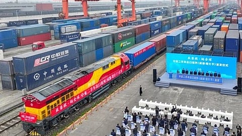 इस तस्वीर में लाल और पीले रंग का मालगाड़ी इंजन खड़ा है, जिसके पीछे कई रंगों के कंटेनर लगे हुए हैं।