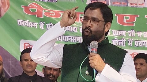 हरे पोस्टर के सामने मंच पर माइक पकड़कर भाषण देते हुए नेता शौकत अली, साथ में हाथ उठाकर इशारा करते हुए।