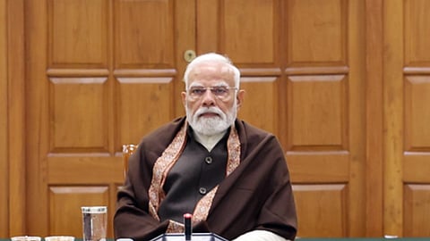 प्रधानमंत्री नरेंद्र मोदी लकड़ी की पैनल वाली पृष्ठभूमि के सामने बैठक में बैठे हुए, भूरे शॉल और पारंपरिक पोशाक में, सामने टेबल पर माइक्रोफोन और पानी का गिलास रखा है।