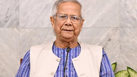 मोहम्मद यूनुस चश्मा लगाए माइक्रोफोन के सामने बैठे हुए, नीले कुर्ते और हल्के रंग की जैकेट में, पीछे संगमरमर की दीवार दिखाई दे रही है।