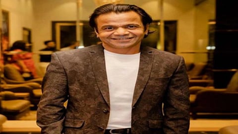 राजपाल यादव अभिनेता भूरे रंग का ब्लेज़र और सफेद टी-शर्ट पहने हुए इनडोर सेटिंग में मुस्कुराते हुए कैमरे की ओर पोज देते नजर आ रहे हैं।