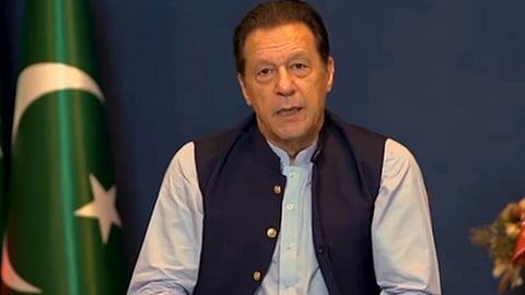 पाकिस्तान के पूर्व कप्तान इमरान खान सामने की ओर देखते हुए और कुर्ता और जैकेट पहने हुए हैं। पीछे पाकिस्तान का झंडा दिख रहा है।