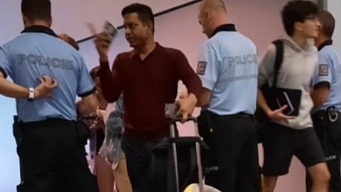 हवाईअड्डे पर पुलिस अधिकारियों से घिरा एक व्यक्ति हाथ में पासपोर्ट और बोर्डिंग पास लिए खड़ा है, उसके पास ट्रॉली बैग है जबकि आसपास यात्री और सुरक्षा कर्मी नजर आ रहे हैं।