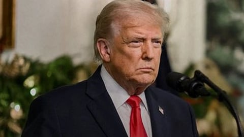 नीले रंग के सूट में अमेरिका के राष्ट्रपति डोनाल्ड ट्रंप जो माइक के सामने खड़े होकर कुछ बोलते दिखाई दे रहे हैं।