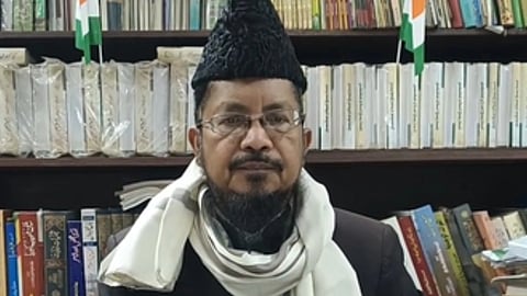 पुस्तकों से भरी अलमारी के सामने बैठा एक व्यक्ति पारंपरिक टोपी और चश्मा पहने हुए दिखाई दे रहा है।