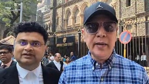 मुंबई हाईकोर्ट परिसर के बाहर खड़े दो पुरुष, सामने खड़ा व्यक्ति कैप और सनग्लासेस पहने कैमरे से बात करता हुआ दिखाई दे रहा है, जबकि पीछे लोग आते-जाते नजर आ रहे हैं।