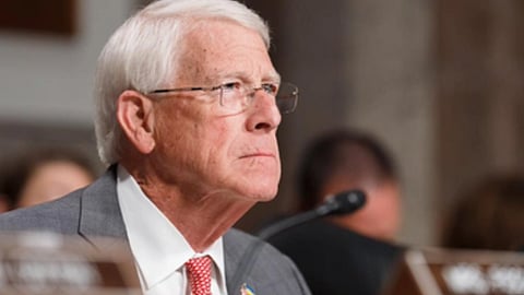 सफेद बाल और चश्मा पहने एक बुजुर्ग व्यक्ति औपचारिक बैठक के दौरान माइक्रोफोन के सामने बैठे नजर आ रहे हैं।