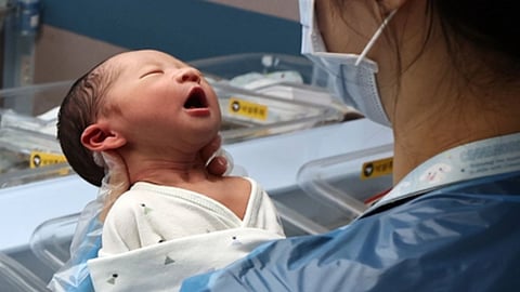 अस्पताल में एक नर्स मास्क पहने नवजात शिशु को गोद में संभालते हुए दिखाई दे रही है।