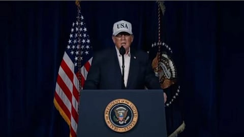 USA लिखी टोपी पहने एक व्यक्ति मंच पर पोडियम से भाषण दे रहा है, पीछे अमेरिकी झंडे लगे हैं।