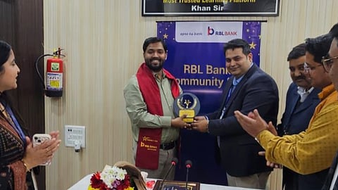 एक कार्यक्रम में मंच पर एक व्यक्ति को ट्रॉफी देकर सम्मानित किया जा रहा है, पीछे RBL Bank का बैनर लगा है और आसपास लोग तालियां बजा रहे हैं।