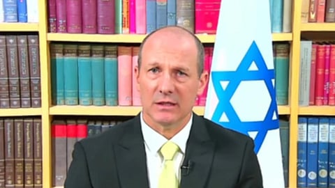 इजरायल के झंडे के सामने किताबों की अलमारी की पृष्ठभूमि में सूट और टाई पहने एक पुरुष आधिकारिक बयान देते हुए दिखाई दे रहे हैं।