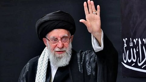 एक बुज़ुर्ग व्यक्ति काली पगड़ी और चोगा पहने हुए हाथ उठाकर अभिवादन करते दिखाई दे रहे हैं, उनके पीछे काले रंग की पृष्ठभूमि पर अरबी लिखावट नजर आ रही है।