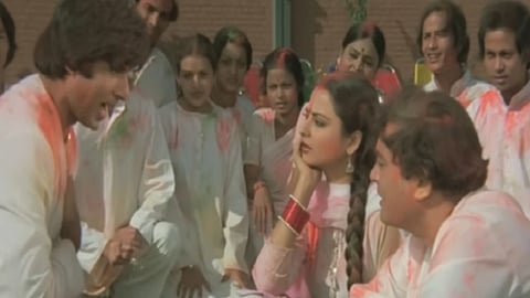 रंगों से सने कपड़ों में कुछ लोग होली के माहौल में बैठे बातचीत कर रहे हैं, आसपास और लोग भी खड़े दिखाई दे रहे हैं।