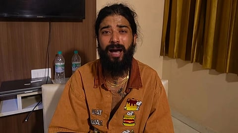 लंबी दाढ़ी वाला एक युवक कमरे में बैठकर कैमरे की ओर बात करता हुआ, पीछे मेज पर पानी की बोतलें और दीवार पर टीवी दिखाई दे रहा है।