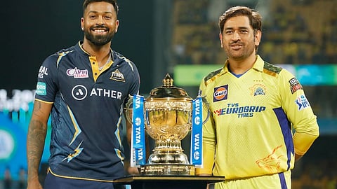 आईपीएल ट्रॉफी के साथ खड़े दो क्रिकेट खिलाड़ी, एक तरफ गुजरात टाइटंस की जर्सी में हार्दिक पांड्या और दूसरी तरफ चेन्नई सुपर किंग्स की जर्सी में महेंद्र सिंह धोनी, मैच से पहले ट्रॉफी के साथ फोटो खिंचवाते हुए।