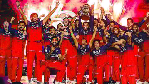 आईपीएल फाइनल जीतने के बाद रॉयल चैलेंजर्स बैंगलोर (RCB) के खिलाड़ी मंच पर ट्रॉफी उठाकर जश्न मनाते हुए, चारों ओर आतिशबाजी और लाल जर्सी में टीम के खिलाड़ी खुशी जाहिर करते नजर आ रहे हैं।