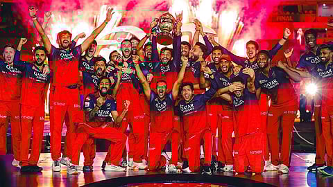 RCB के सभी खिलाड़ी और स्टाफ मैदान पर एक साथ खड़े होकर आईपीएल ट्रॉफी जीतने का जश्न मना रहे हैं। बैकग्राउंड में आतिशबाजी हो रही है और सभी बहुत खुश दिख रहे हैं।