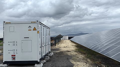 Containerised battery energy storage system installed next to utility-scale solar panels under cloudy skies.