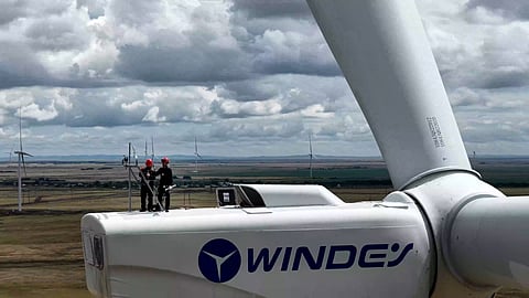 Two technicians wearing safety helmets stand on a Windey wind turbine nacelle during maintenance, with other wind turbines visible across the surrounding landscape.