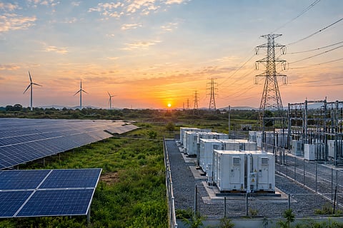 Large solar panels and wind turbines connected to high-voltage transmission towers, substations and battery storage units in a renewable energy grid landscape.