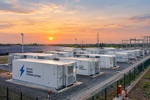 Rows of containerized battery energy storage units installed at a utility-scale facility, with solar panels, a substation and high-voltage transmission infrastructure visible in the background.
