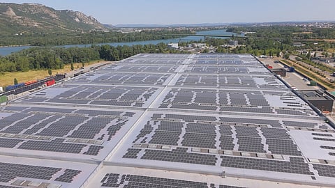 Aerial view of extensive solar panels installed across the rooftop of a large commercial facility, with a river, roads and surrounding landscape visible in the background.