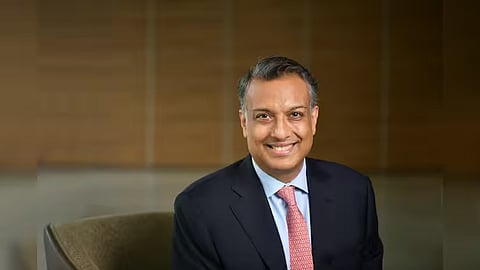 Sumant Sinha sits on a chair wearing a dark suit, light blue shirt and pink tie against a warm brown background.