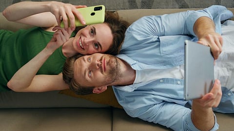 A couple relaxing on a couch, taking a selfie while browsing on a tablet
