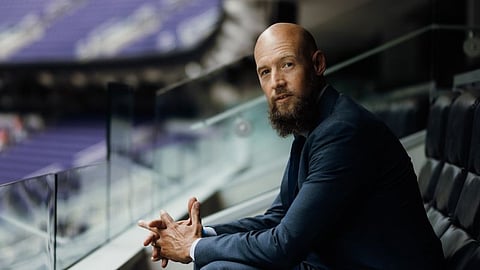 Russell Nicolet seated in a stadium-style setting, wearing a suit and looking toward the camera