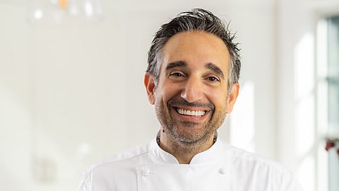 Chef Nicholas Poulmentis smiling in a white chef’s coat inside a bright kitchen setting