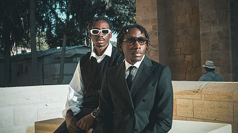 Two men in tailored suits and sunglasses pose outdoors in a modern formal fashion look