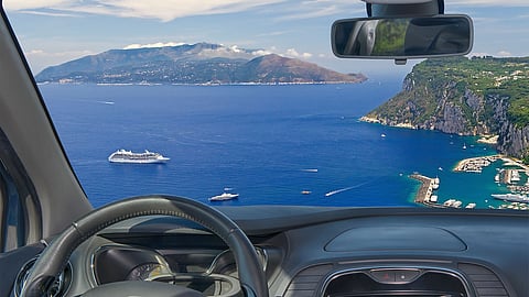 View from inside a car overlooking a blue Greek coastline with cliffs, boats, and distant islands