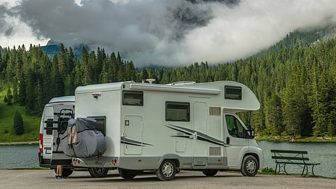 White RV parked by forested lake with mountains and low clouds in background