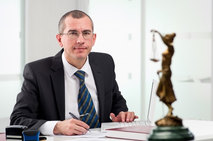 Man in suit sitting at desk with legal documents and scales of justice in foreground
