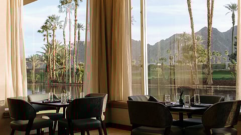 Interior dining space with tables and chairs overlooking palm trees, water, and mountain views