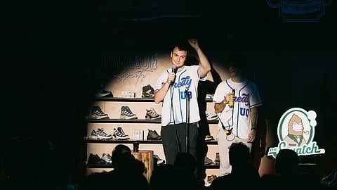 Stand-up comedians perform on stage inside a retail store with audience watching
