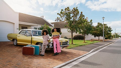 Two children standing beside a vintage car with suitcases in a suburban driveway