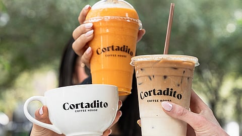 Hands holding coffee, iced latte, and orange smoothie drinks with Cortadito branding