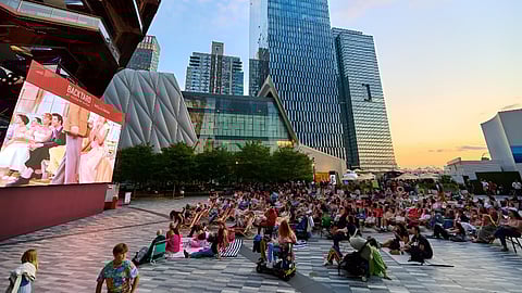 Large audience seated outdoors at Hudson Yards plaza watching a big screen with skyline at sunset