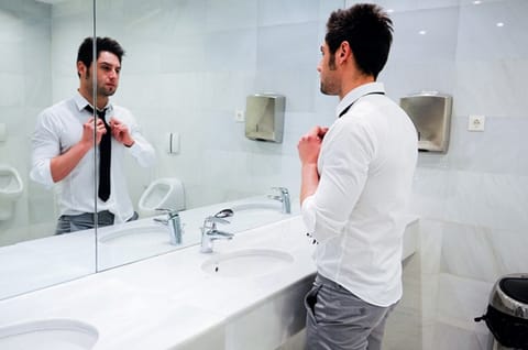 A man adjusting his tie in front of a mirror within a public restroom.
